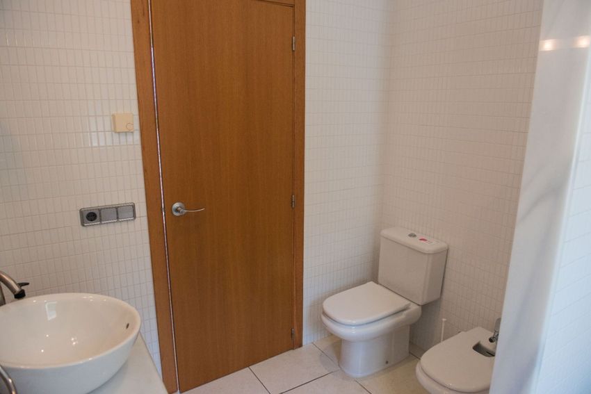 Baño funcional con paredes de azulejos blancos verticales, inodoro, bidé y lavabo sobre encimera. Incluye puerta de madera y suelo de baldosas claras. Baño funcional con paredes de azulejos blancos verticales, inodoro, bidé y lavabo sobre encimera. Incluye puerta de madera y suelo de baldosas claras.