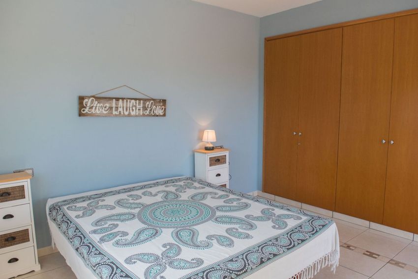 Dormitorio con paredes azul claro, colcha estampada y armarios empotrados de madera. Mesitas de noche blancas con detalles de mimbre y suelo de baldosas. Dormitorio con paredes azul claro, colcha estampada y armarios empotrados de madera. Mesitas de noche blancas con detalles de mimbre y suelo de baldosas.
