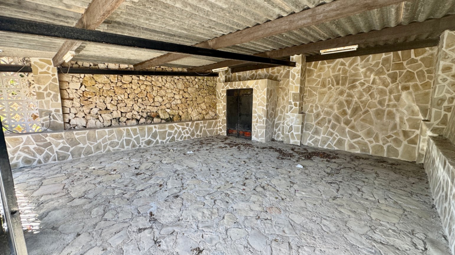 Espacio exterior cubierto con paredes y suelos de piedra irregular, vigas de madera y metal, y una cubierta de chapa ondulada.