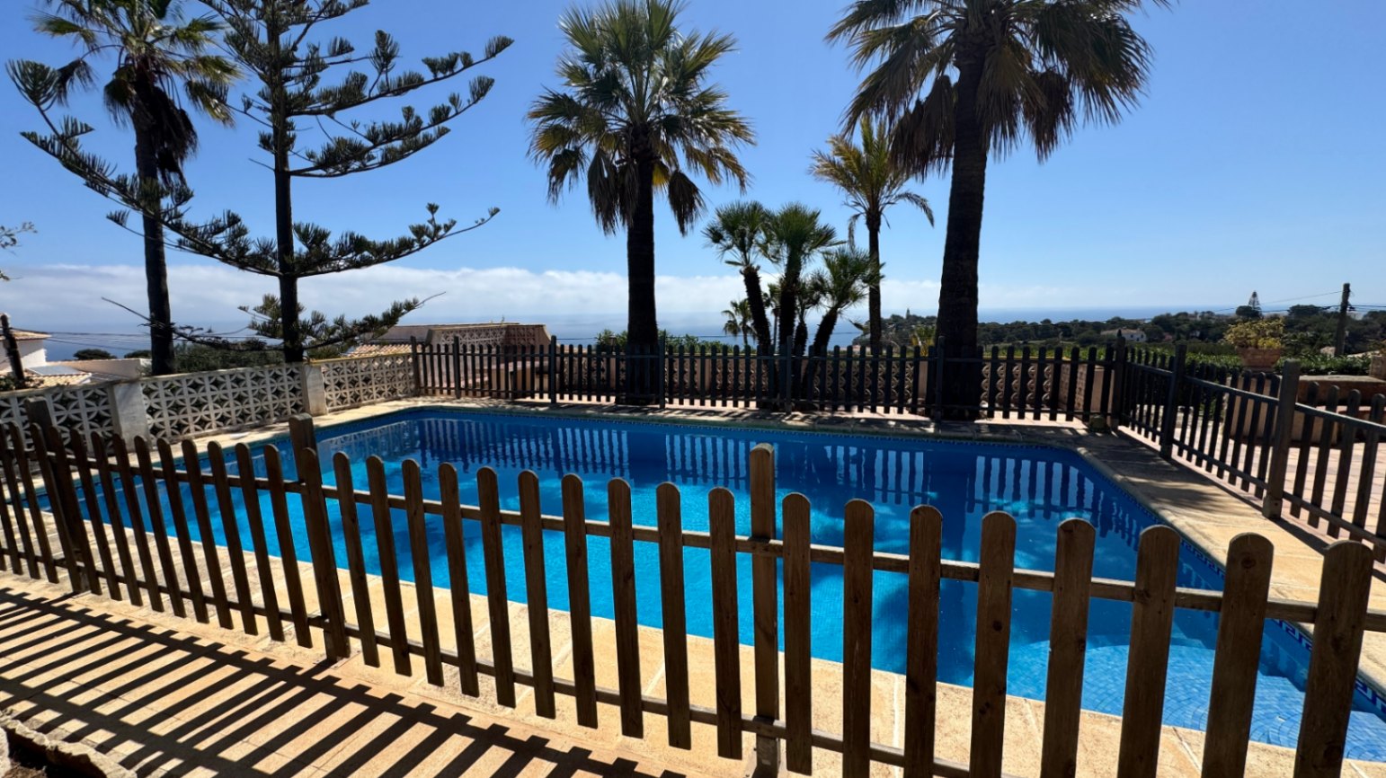 Piscina exterior con azulejos azules y valla perimetral de madera. La zona está rodeada de palmeras y ofrece vistas despejadas hacia la costa mediterránea.