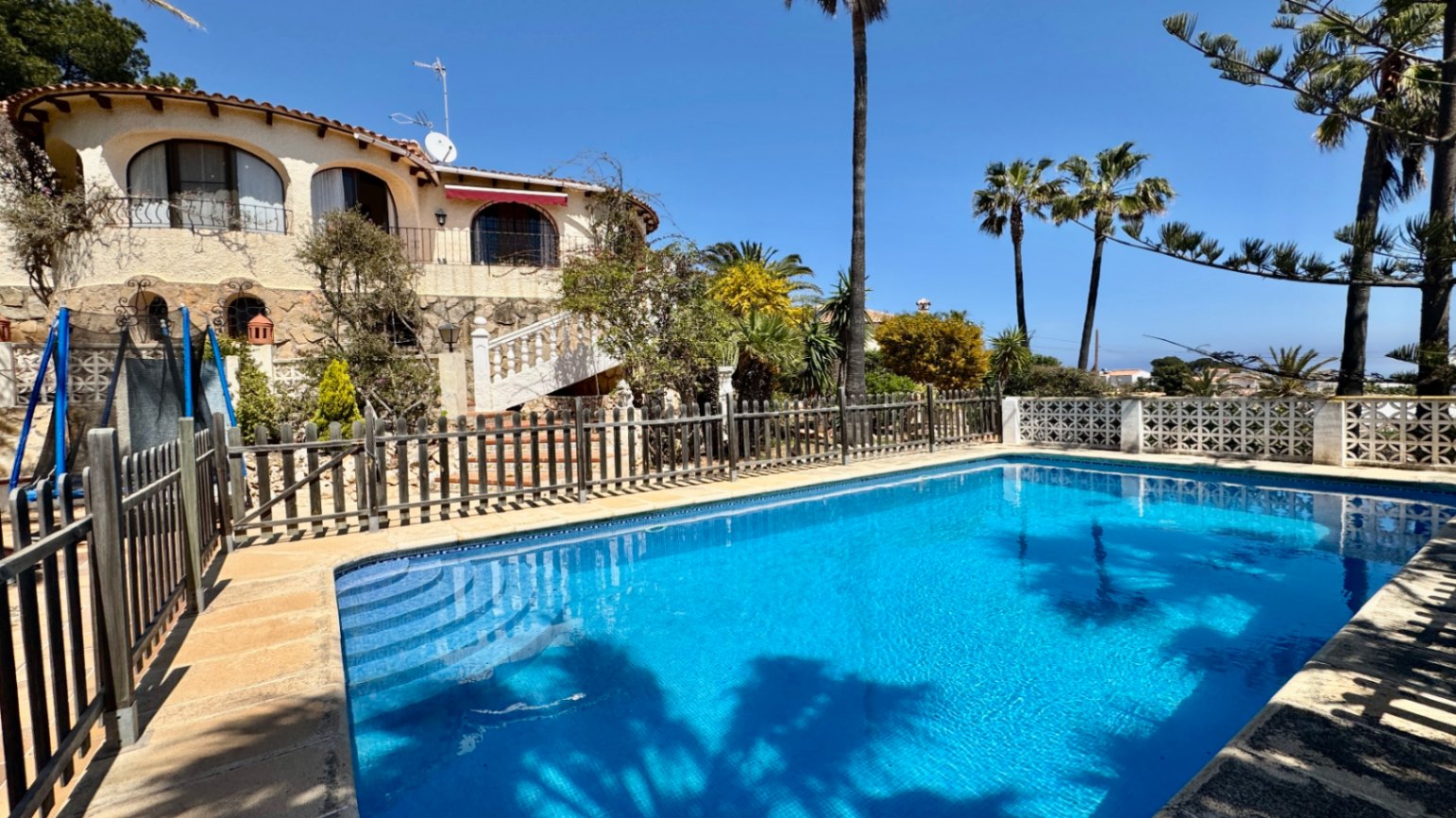 Piscina exterior con revestimiento azul y escaleras integradas, rodeada por una valla de madera y una villa mediterránea con zócalo de piedra y balcones arqueados.