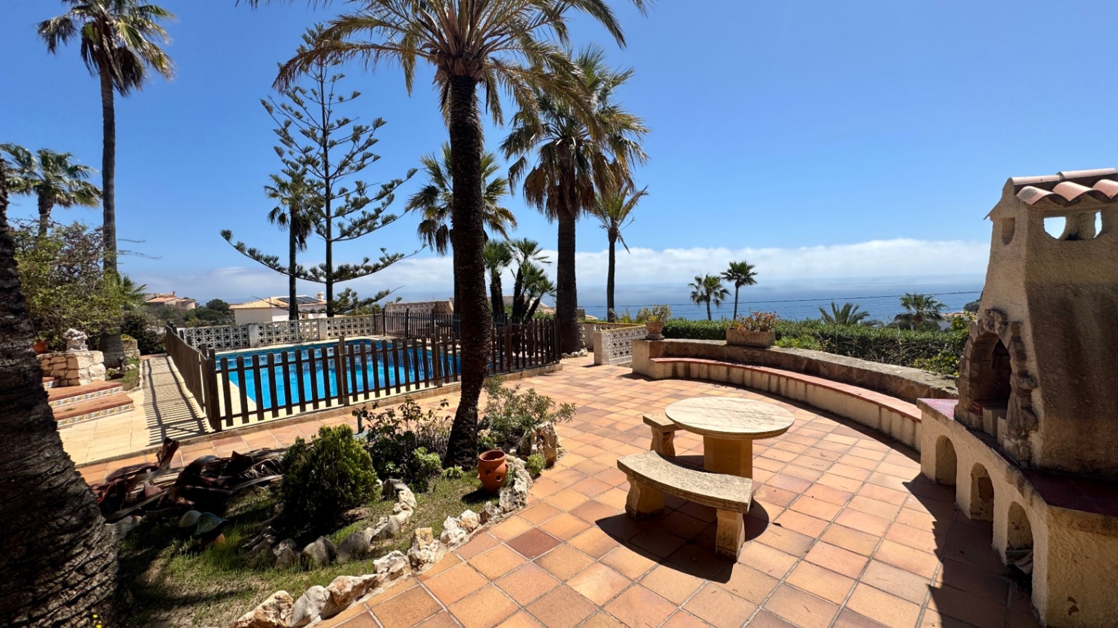 Terraza de baldosas de terracota con mobiliario de piedra, chimenea exterior rústica y piscina vallada con vistas panorámicas al mar y palmeras.