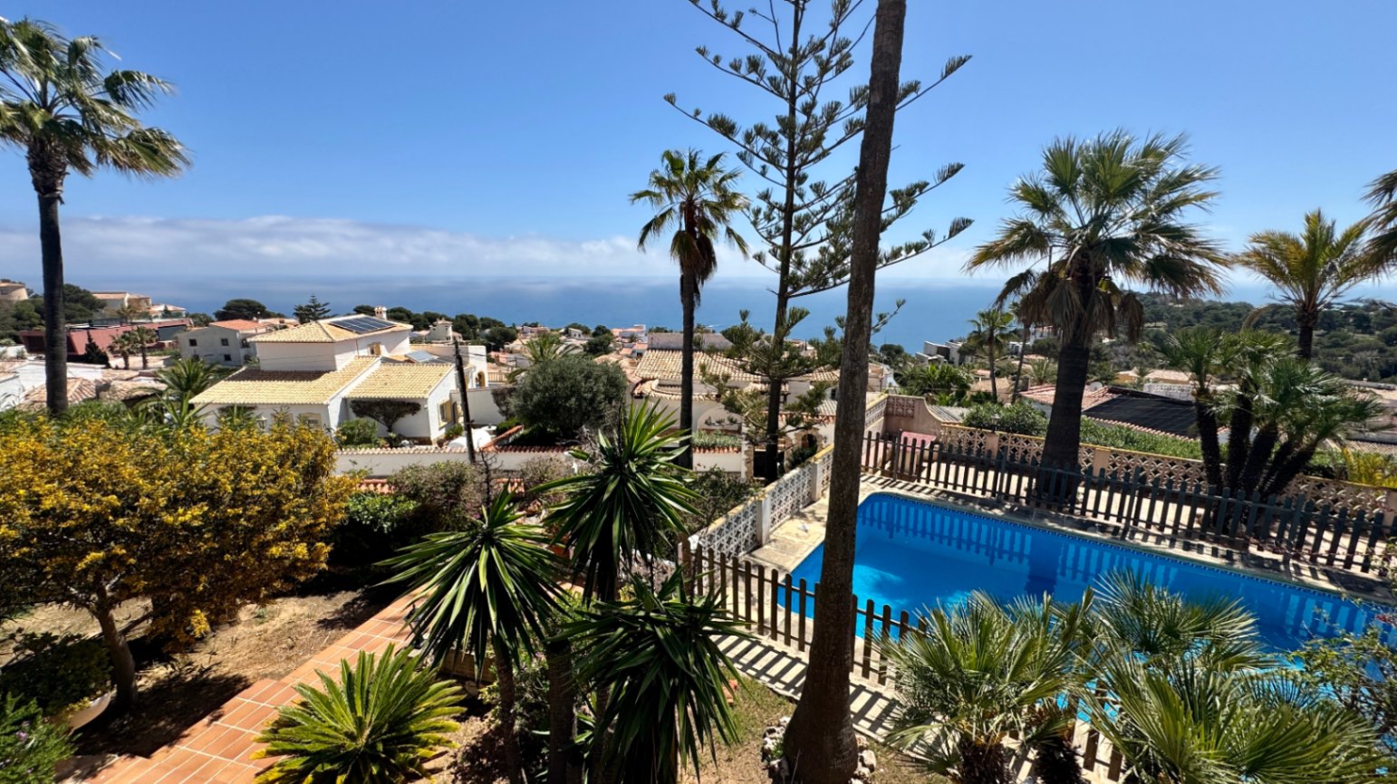 Vista elevada de una piscina privada con valla de madera, rodeada de palmeras y jardines mediterráneos con vistas panorámicas al mar.