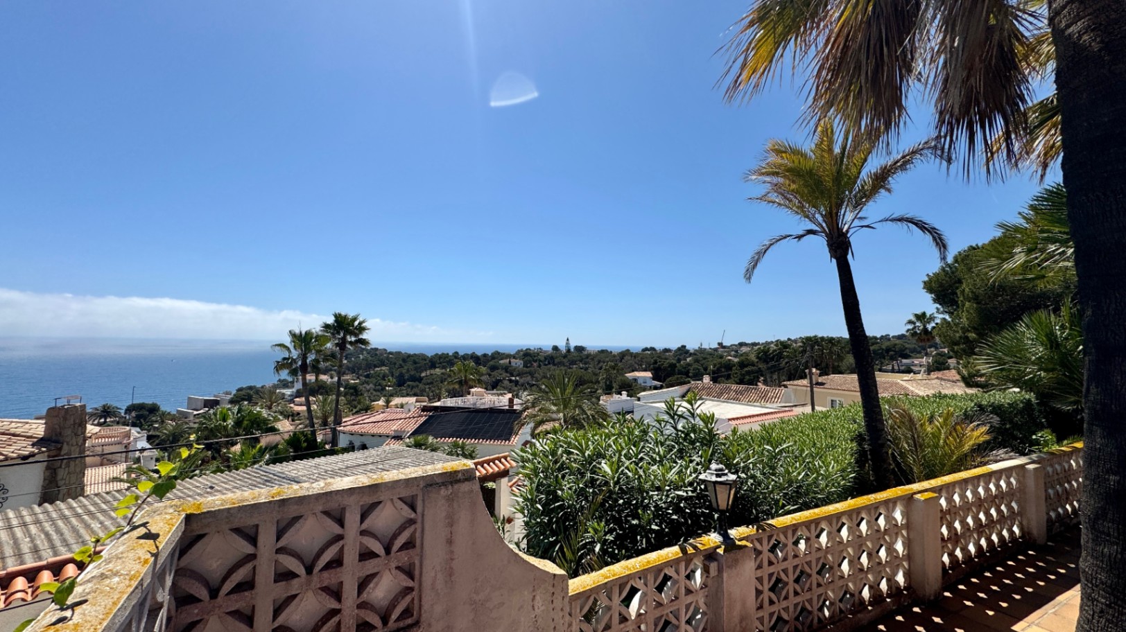 Vista elevada desde una terraza con balaustrada geométrica hacia un entorno mediterráneo de tejados cerámicos, palmeras y el mar en el horizonte.