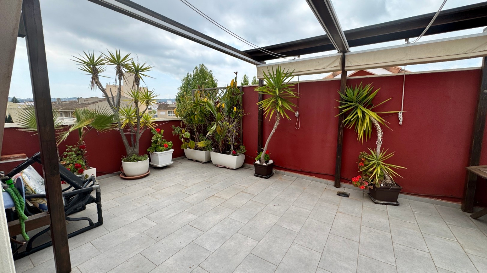 Amplia terraza en la azotea con suelo de baldosas grises, pérgola de madera con toldos y variadas plantas mediterráneas frente a un muro perimetral de color granate.