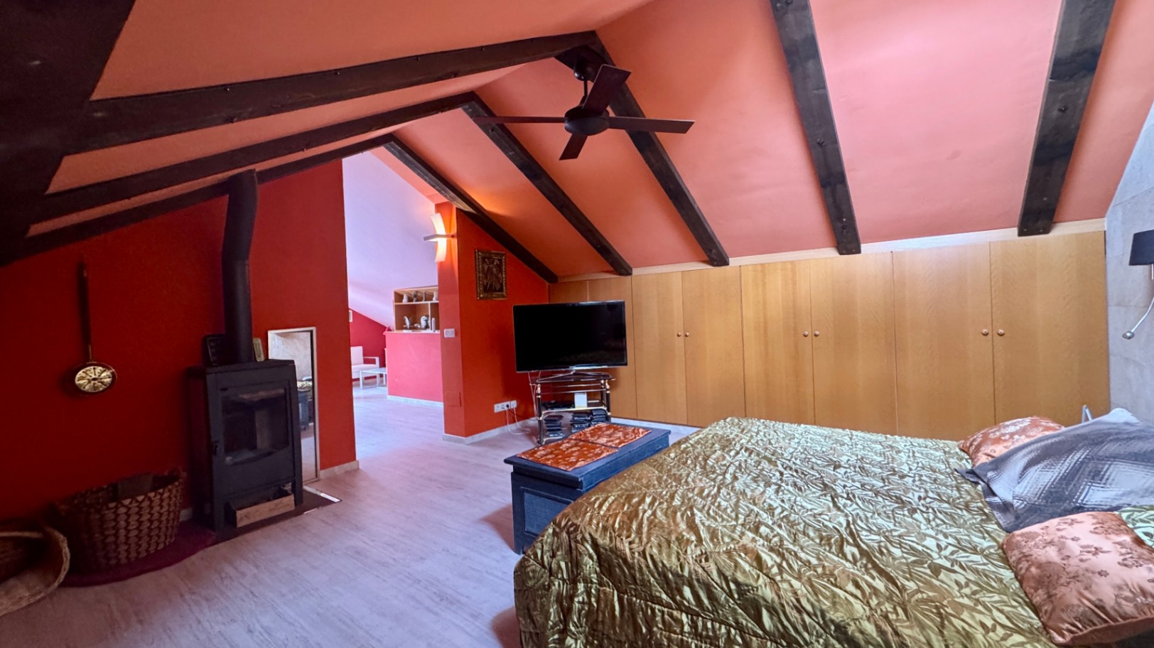 Dormitorio abuhardillado con vigas de madera oscura, paredes terracota, estufa de leña de hierro y amplios armarios empotrados de madera clara.