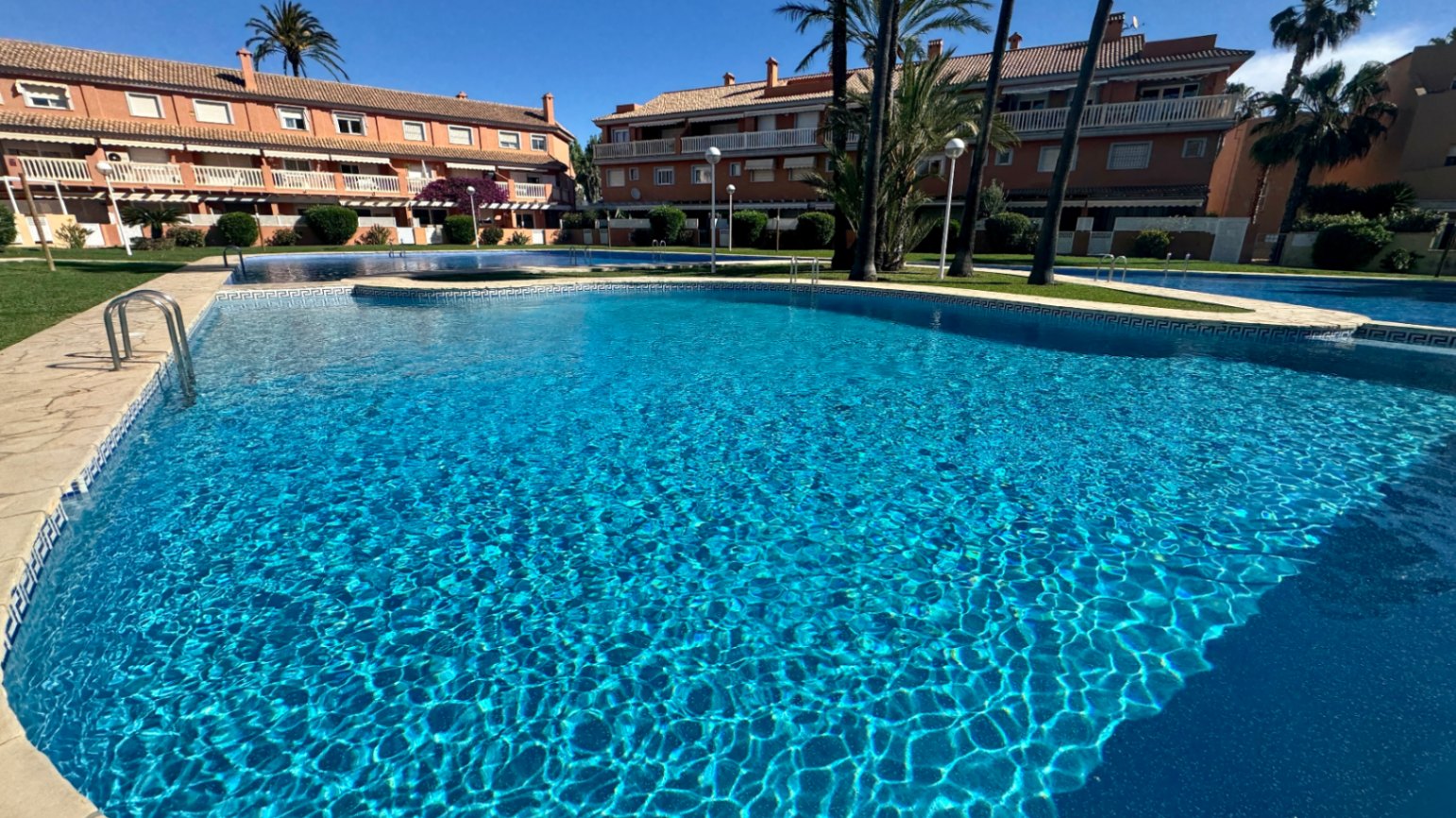Piscina comunitaria con azulejos azules y borde de piedra, rodeada de jardines cuidados y edificios residenciales de estilo mediterráneo.