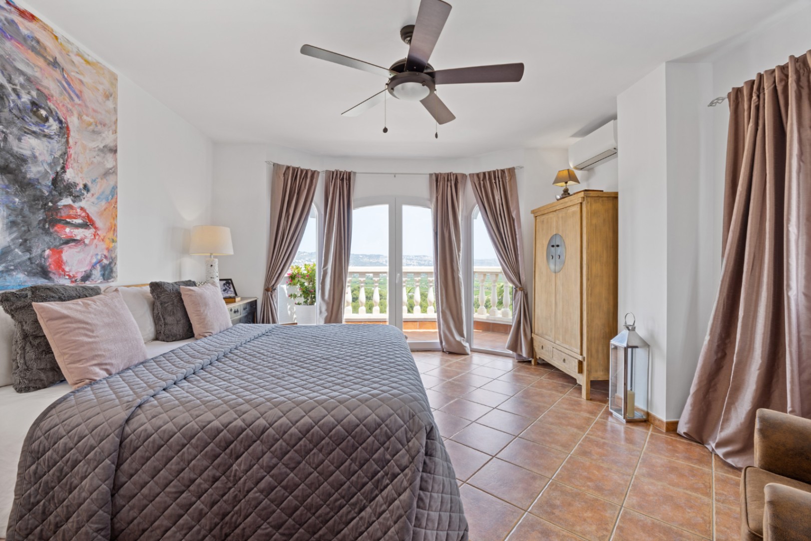 Dormitorio luminoso con suelo de terracota, ventilador de techo y puertas acristaladas en arco que dan acceso a un balcón con vistas despejadas.