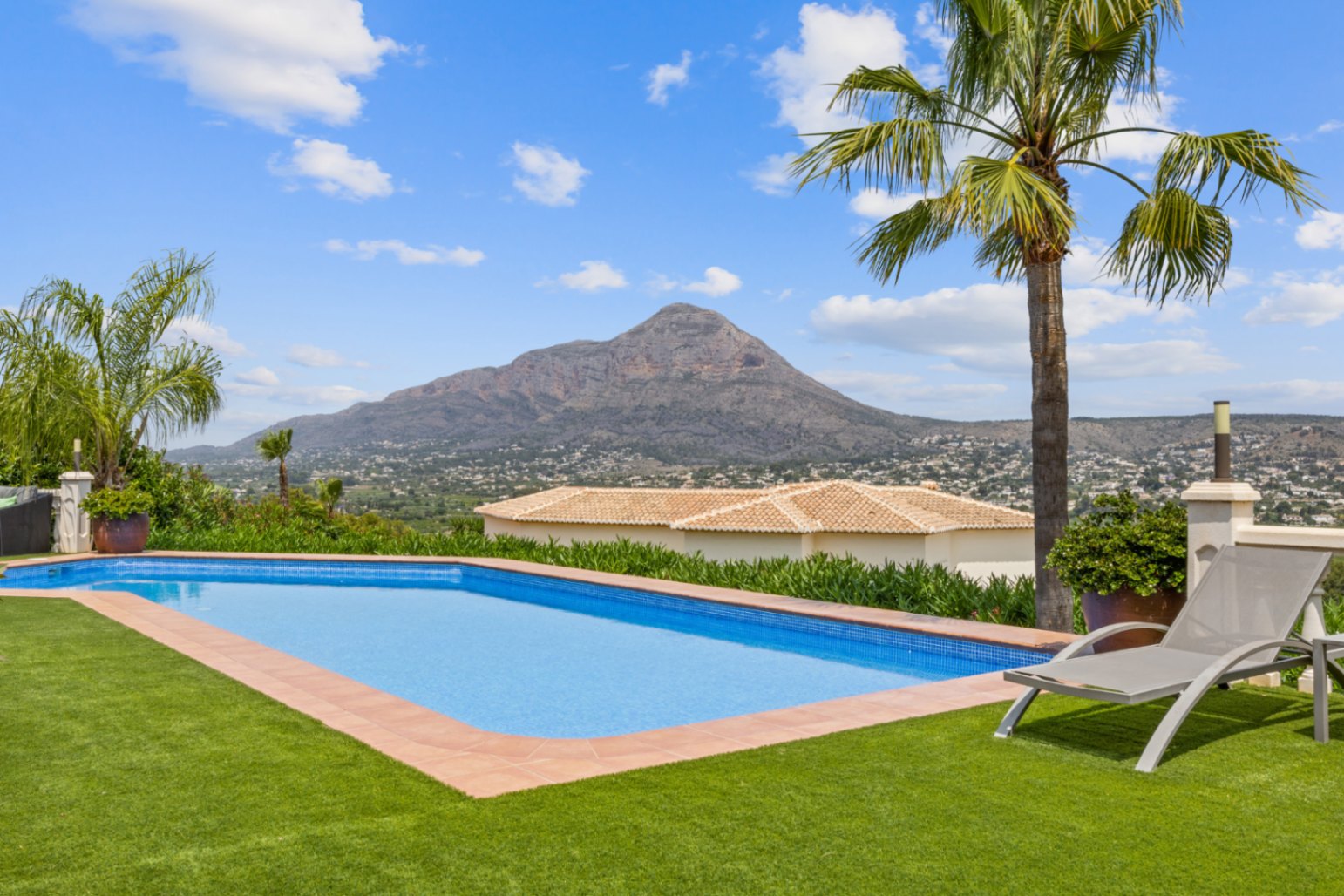 Piscina exterior con revestimiento de gresite azul, rodeada de césped artificial y bordes de terracota, con vistas panorámicas a un pico montañoso.