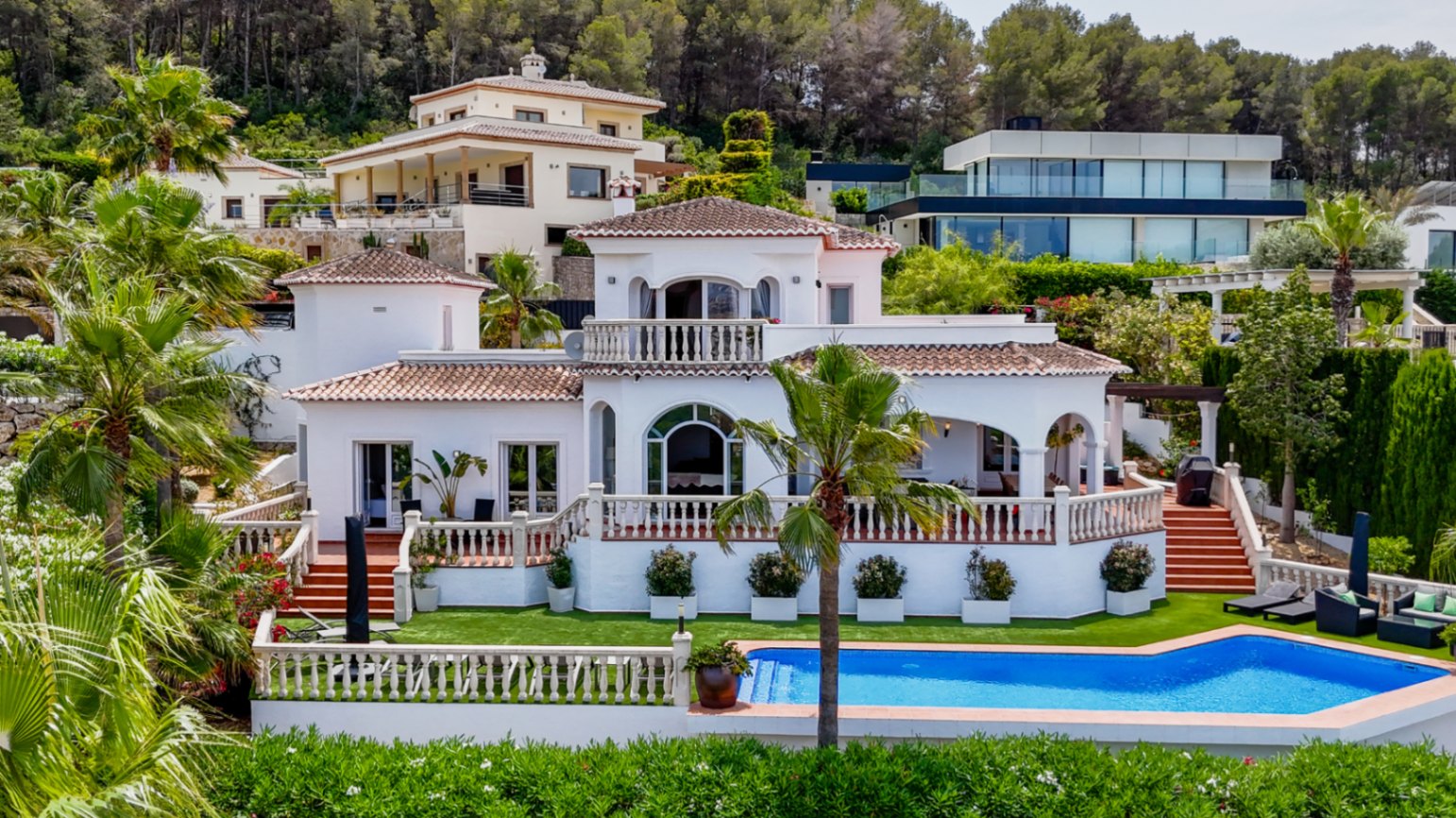 Villa mediterránea con fachada blanca, tejados de terracota y terrazas con balaustradas frente a una piscina privada y jardín con palmeras.