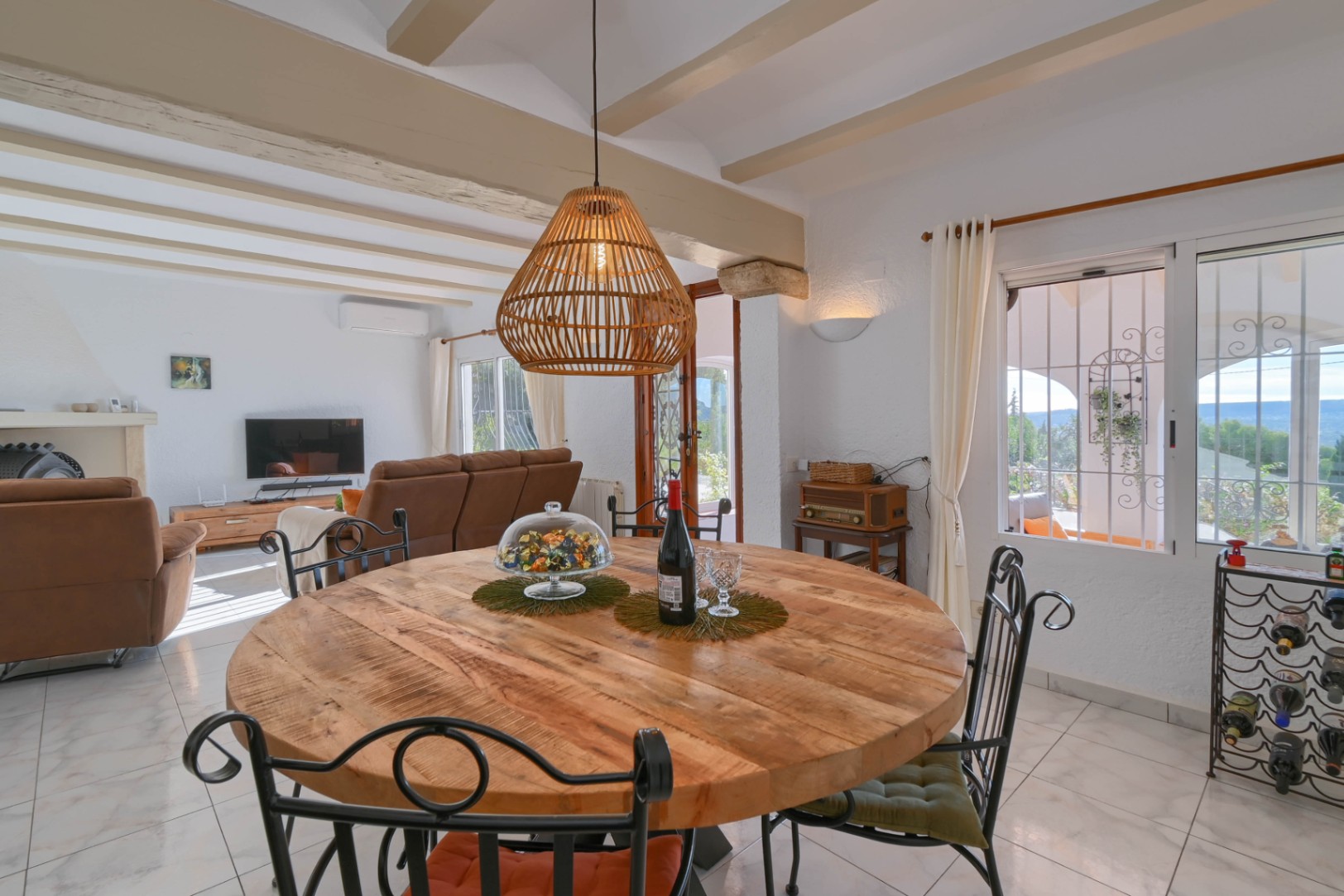 Comedor con mesa redonda de madera, sillas de forja y lámpara de mimbre, bajo un techo de vigas vistas que conecta con el salón principal.