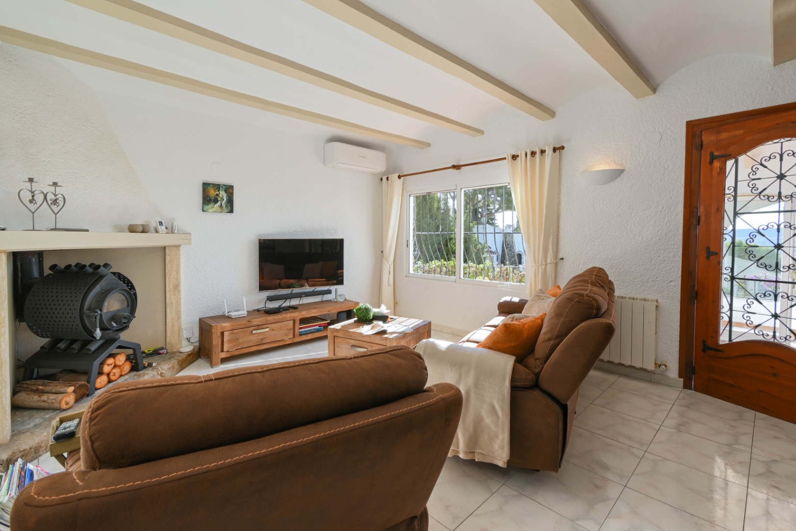 Salón con vigas de madera a la vista, chimenea con estufa de leña y mobiliario rústico. Incluye suelos cerámicos y luz natural desde el ventanal y la puerta decorativa de hierro.