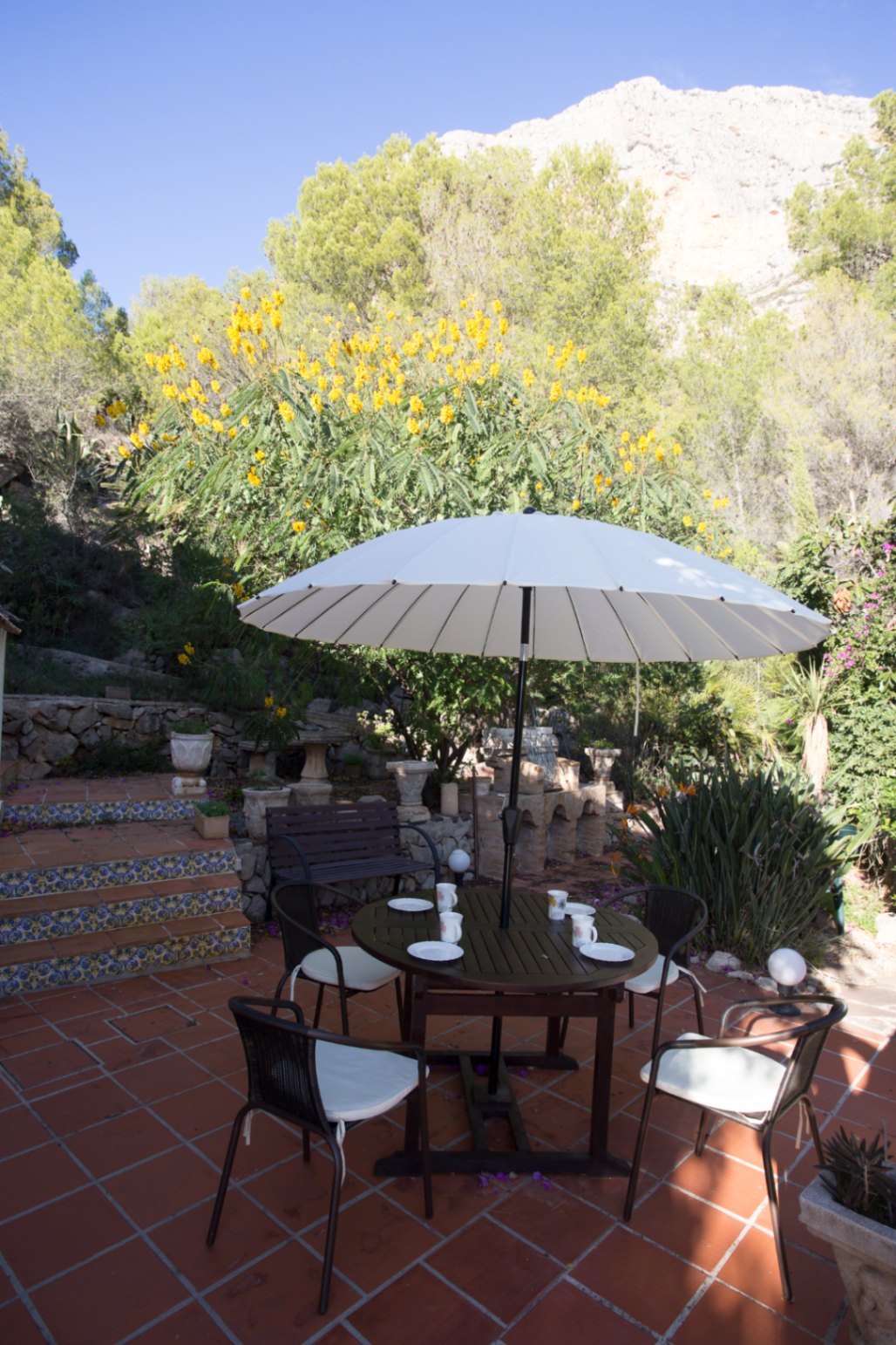 Terraza exterior con suelo de terracota, mesa de comedor bajo sombrilla blanca y escalones decorados con azulejos, rodeada de vegetación y vistas a la montaña.