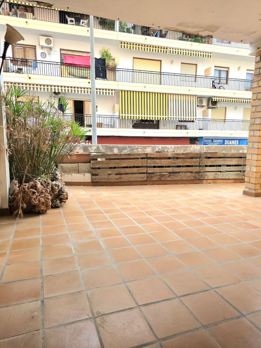 Amplia terraza exterior con suelo de baldosas de terracota, delimitada por una jardinera de madera y vegetación. Ofrece vistas a un edificio residencial con balcones y toldos a rayas. Amplia terraza exterior con suelo de baldosas de terracota, delimitada por una jardinera de madera y vegetación. Ofrece vistas a un edificio residencial con balcones y toldos a rayas.