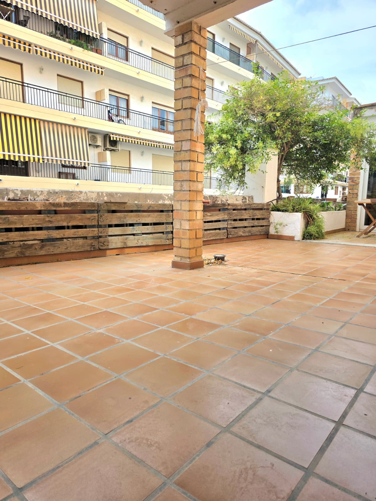 Amplia terraza exterior con suelo de baldosas de terracota, un pilar central de ladrillo visto y cerramientos de madera tipo palet con vegetación integrada.