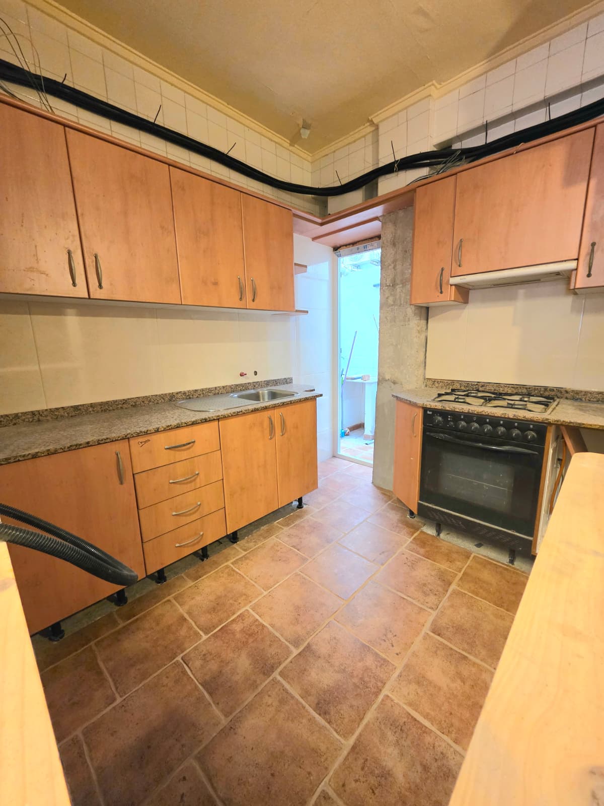Cocina con armarios de acabado madera, encimera de granito y suelo de gres rústico. Incluye horno negro, placa de gas y acceso directo a una zona de lavadero exterior.