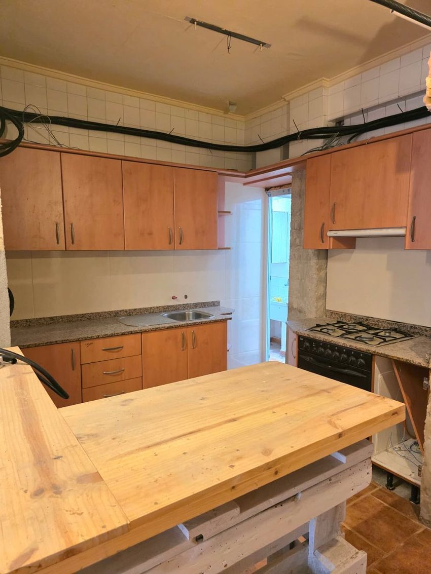 Cocina en fase de actualización con muebles de madera clara, encimeras de granito y fregadero doble. Destaca la placa de gas integrada, el cableado expuesto y una barra de madera sin terminar en primer plano. Cocina en fase de actualización con muebles de madera clara, encimeras de granito y fregadero doble. Destaca la placa de gas integrada, el cableado expuesto y una barra de madera sin terminar en primer plano.