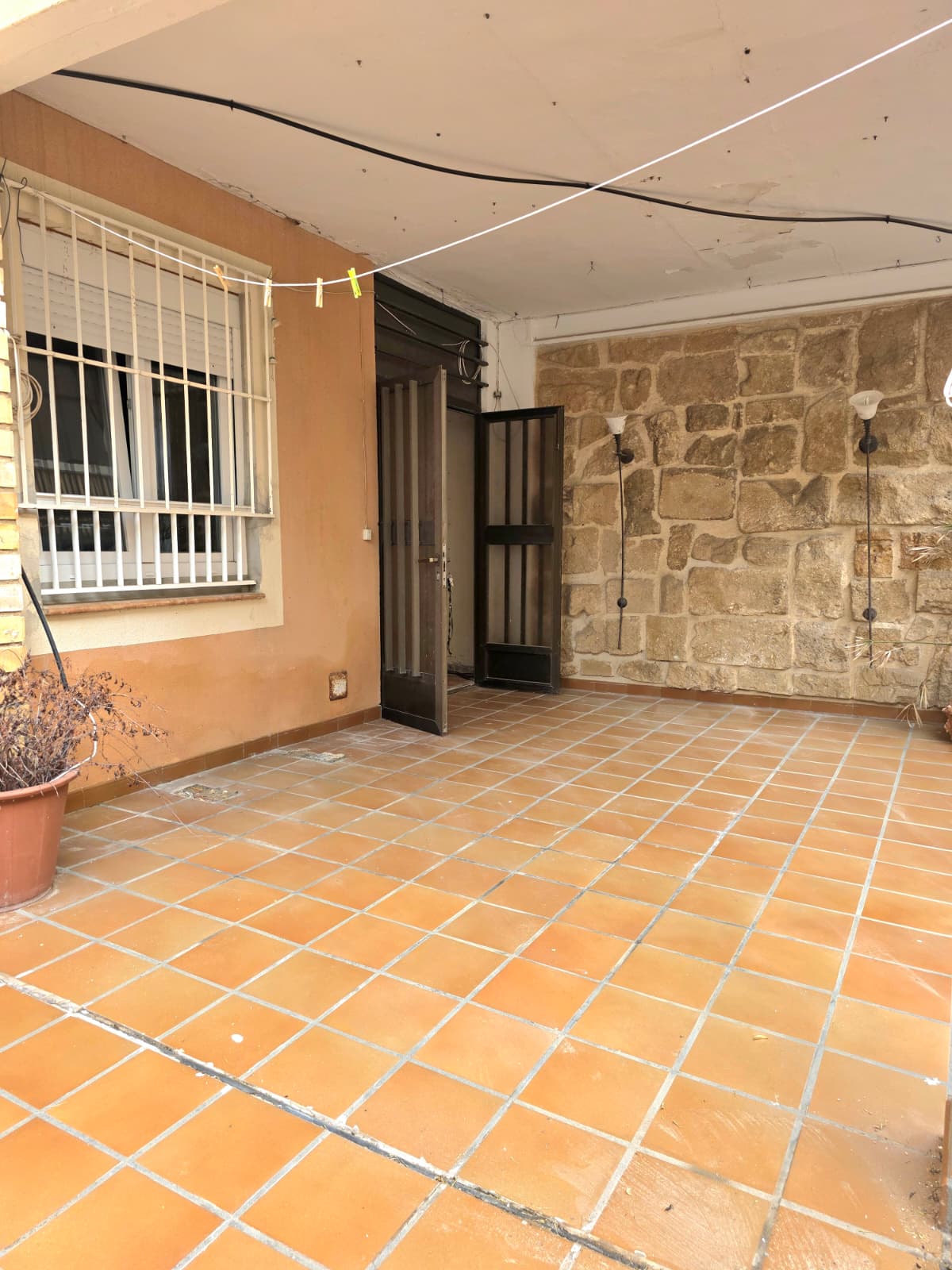 Patio cubierto con suelo de baldosas de terracota, una pared de piedra rústica con apliques y ventana protegida por rejas de seguridad blancas.