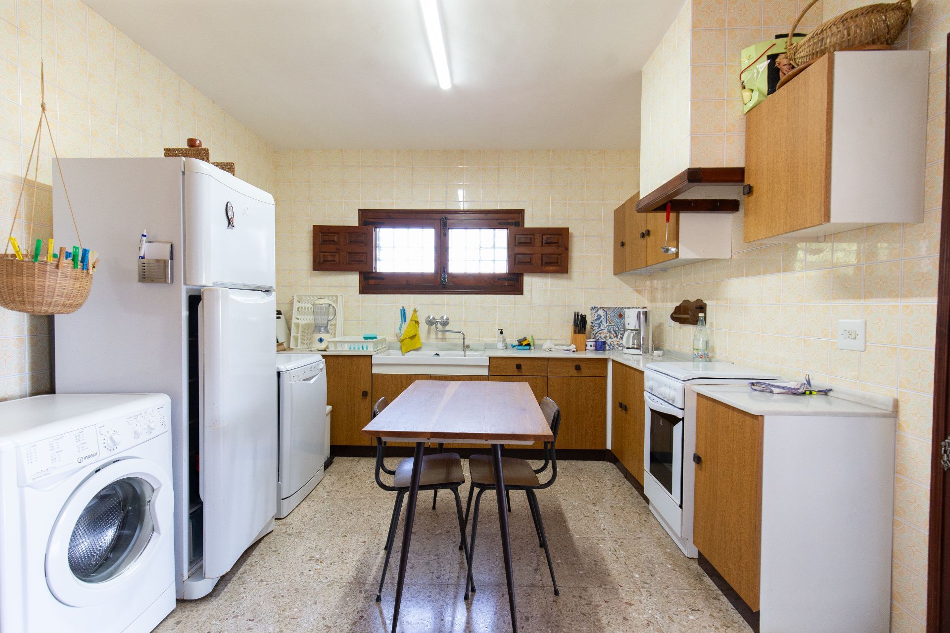 Cocina funcional con armarios de madera, azulejos estampados y electrodomésticos blancos. Incluye una mesa de comedor central sobre suelo de terrazo y ventanas con contraventanas de madera.