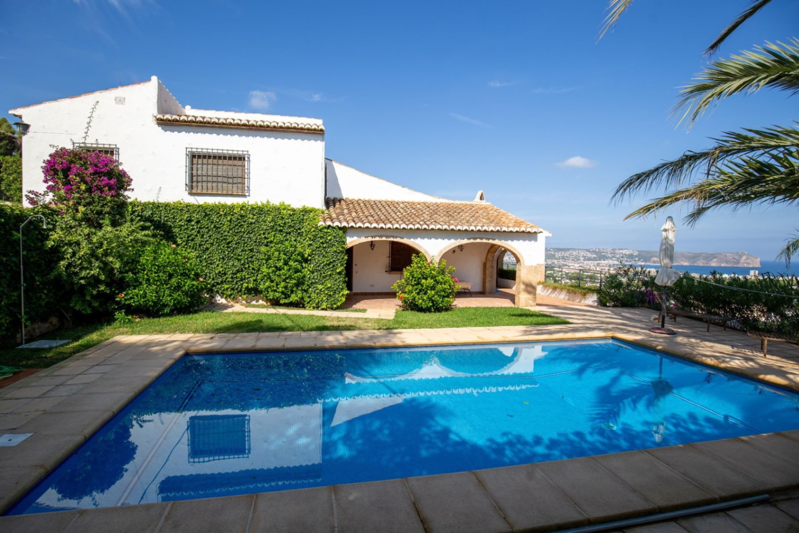 Exterior con piscina rectangular, porche de arcos en piedra tosca y fachada blanca con buganvillas, ofreciendo vistas panorámicas al mar y la costa.
