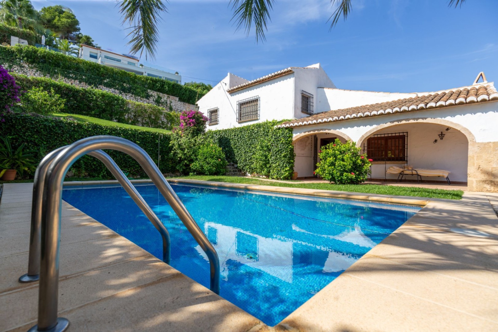 Piscina rectangular con gresite azul y escalera de acero, junto a una villa blanca tradicional con porches de arcos de medio punto y tejado de teja cerámica.