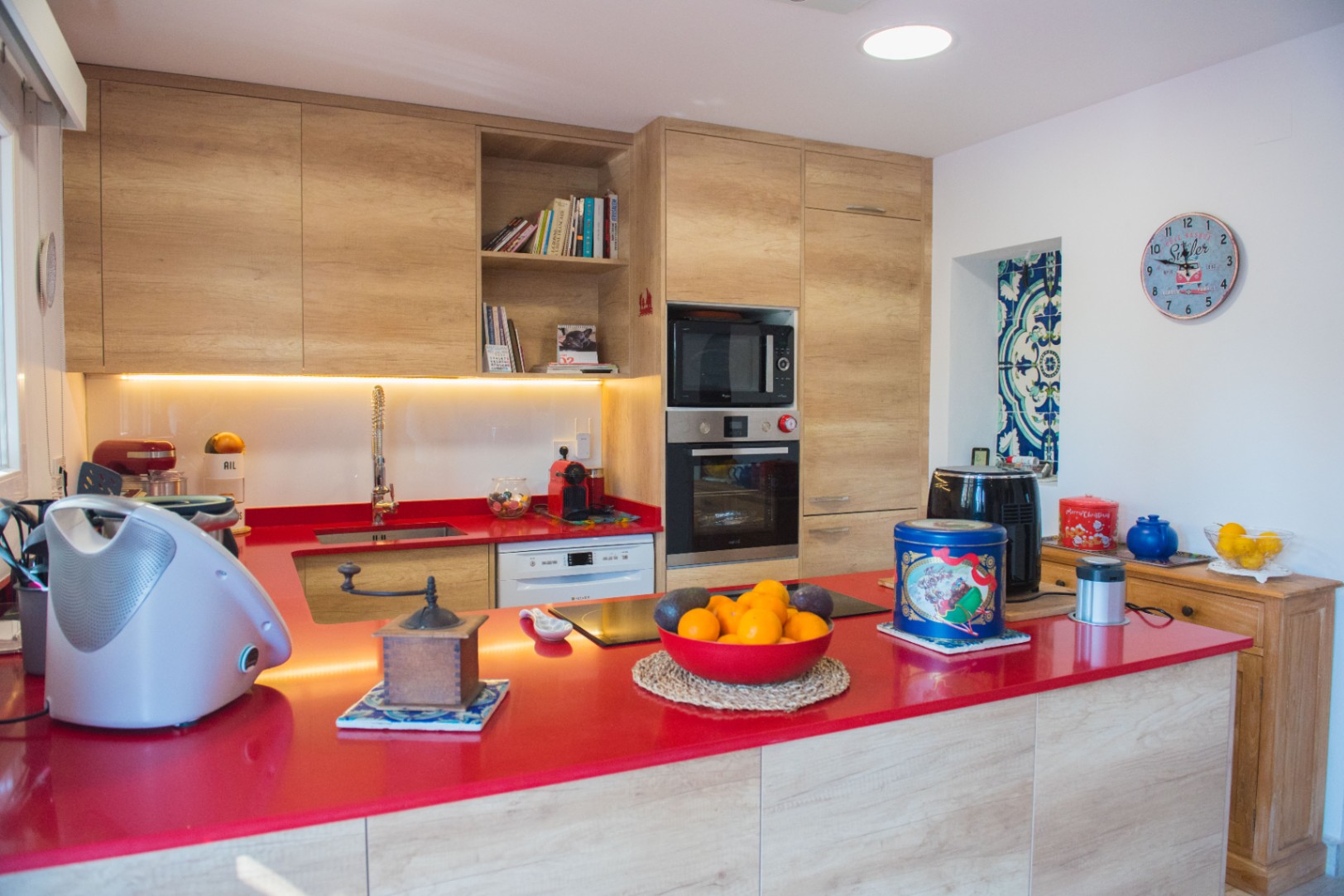 Cocina moderna con encimera roja vibrante, armarios de madera clara y electrodomésticos integrados, incluyendo horno y placa de inducción.