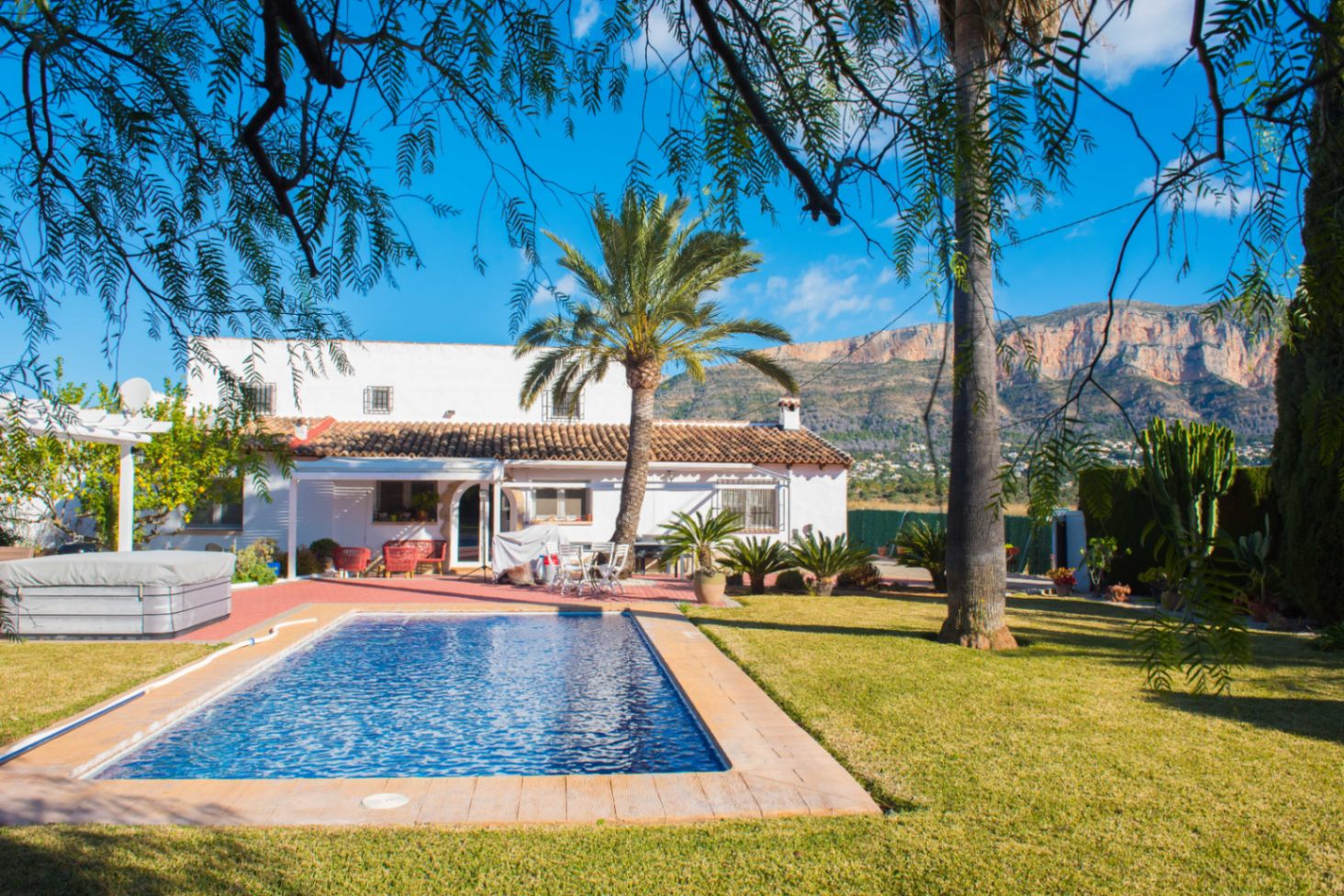 Zona exterior con piscina rectangular, césped y palmeras frente al macizo del Montgó. La villa blanca destaca por su tejado de arcilla y un jacuzzi integrado en la terraza.
