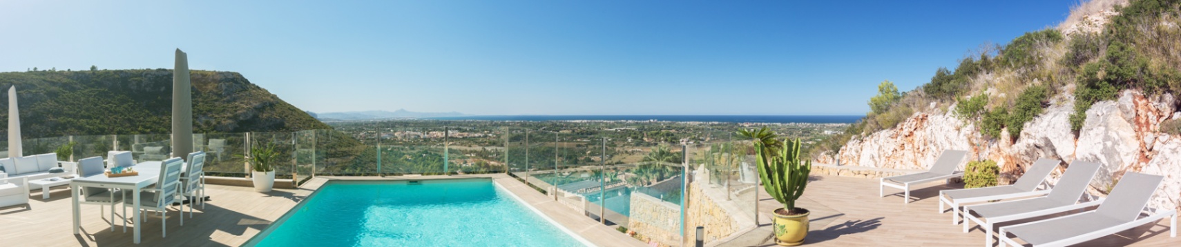 Amplia terraza exterior con piscina rectangular, mobiliario moderno blanco para comedor y salón, y tumbonas. Vistas panorámicas a la costa, el mar y las colinas verdes bajo un cielo despejado. Amplia terraza exterior con piscina rectangular, mobiliario moderno blanco para comedor y salón, y tumbonas. Vistas panorámicas a la costa, el mar y las colinas verdes bajo un cielo despejado.