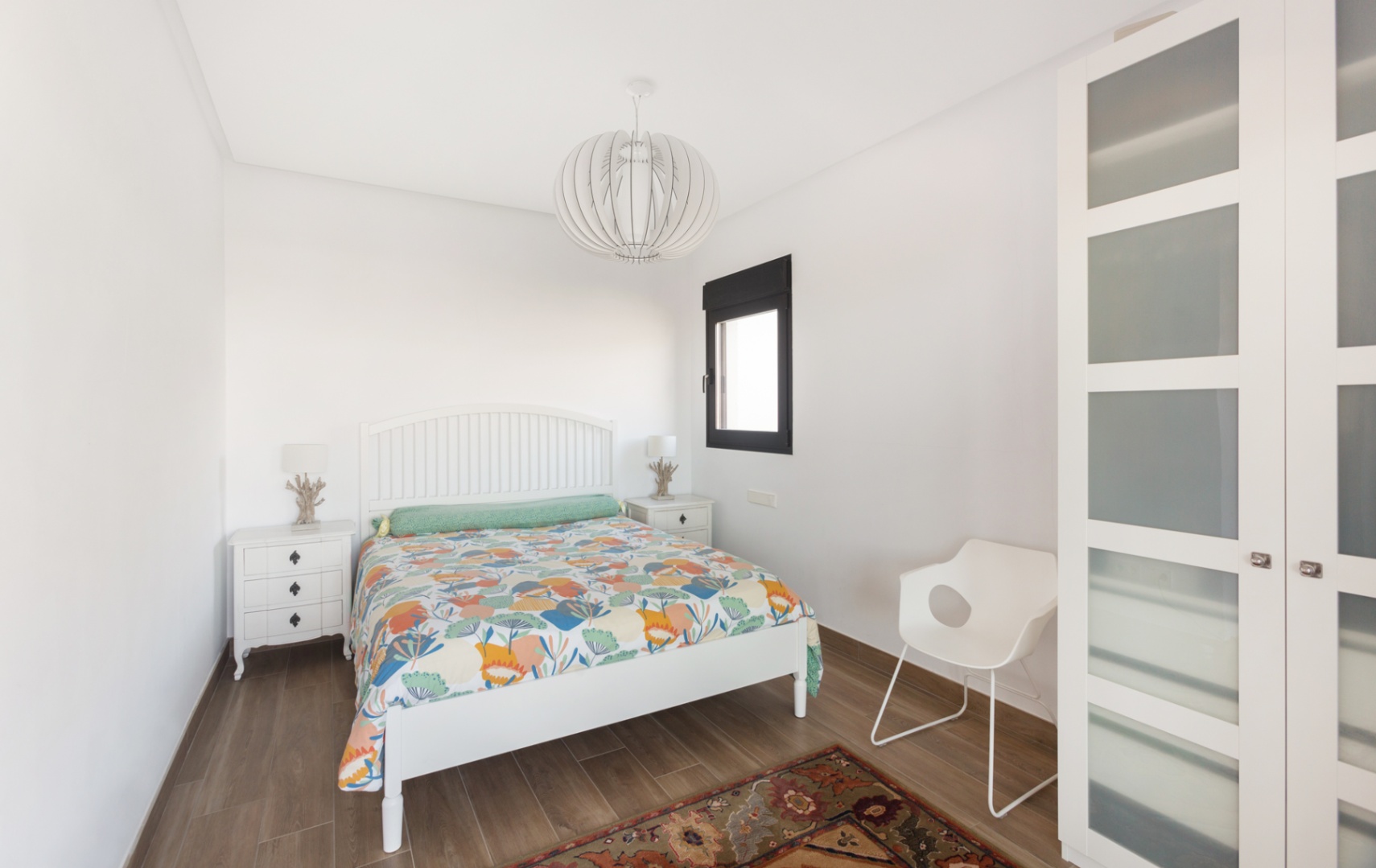 Dormitorio con cabecero de lamas blanco, edredón colorido y suelo de baldosas imitación madera. Incluye mesitas de noche blancas con lámparas decorativas, ventana de marco oscuro y armario con paneles de cristal esmerilado. Dormitorio con cabecero de lamas blanco, edredón colorido y suelo de baldosas imitación madera. Incluye mesitas de noche blancas con lámparas decorativas, ventana de marco oscuro y armario con paneles de cristal esmerilado.