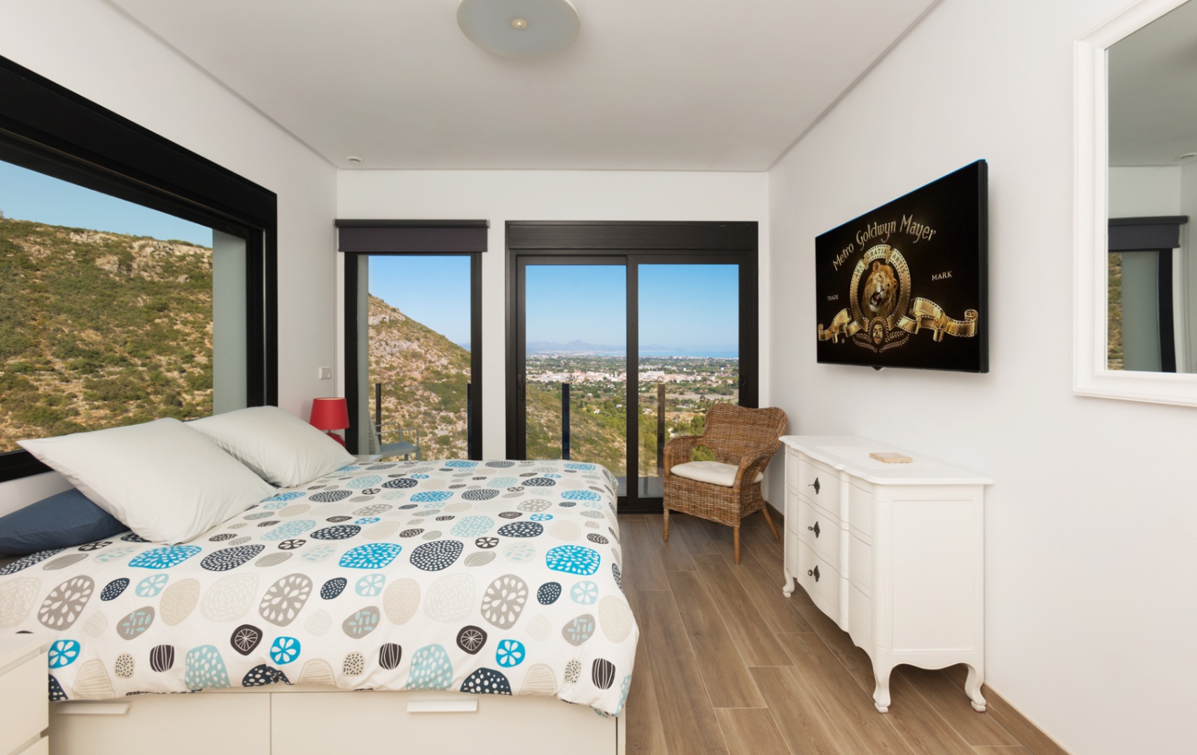 Dormitorio luminoso con cama de diseño, suelo de madera y amplios ventanales negros que enmarcan vistas panorámicas a la montaña y la costa. Incluye cómoda blanca y TV. Dormitorio luminoso con cama de diseño, suelo de madera y amplios ventanales negros que enmarcan vistas panorámicas a la montaña y la costa. Incluye cómoda blanca y TV.
