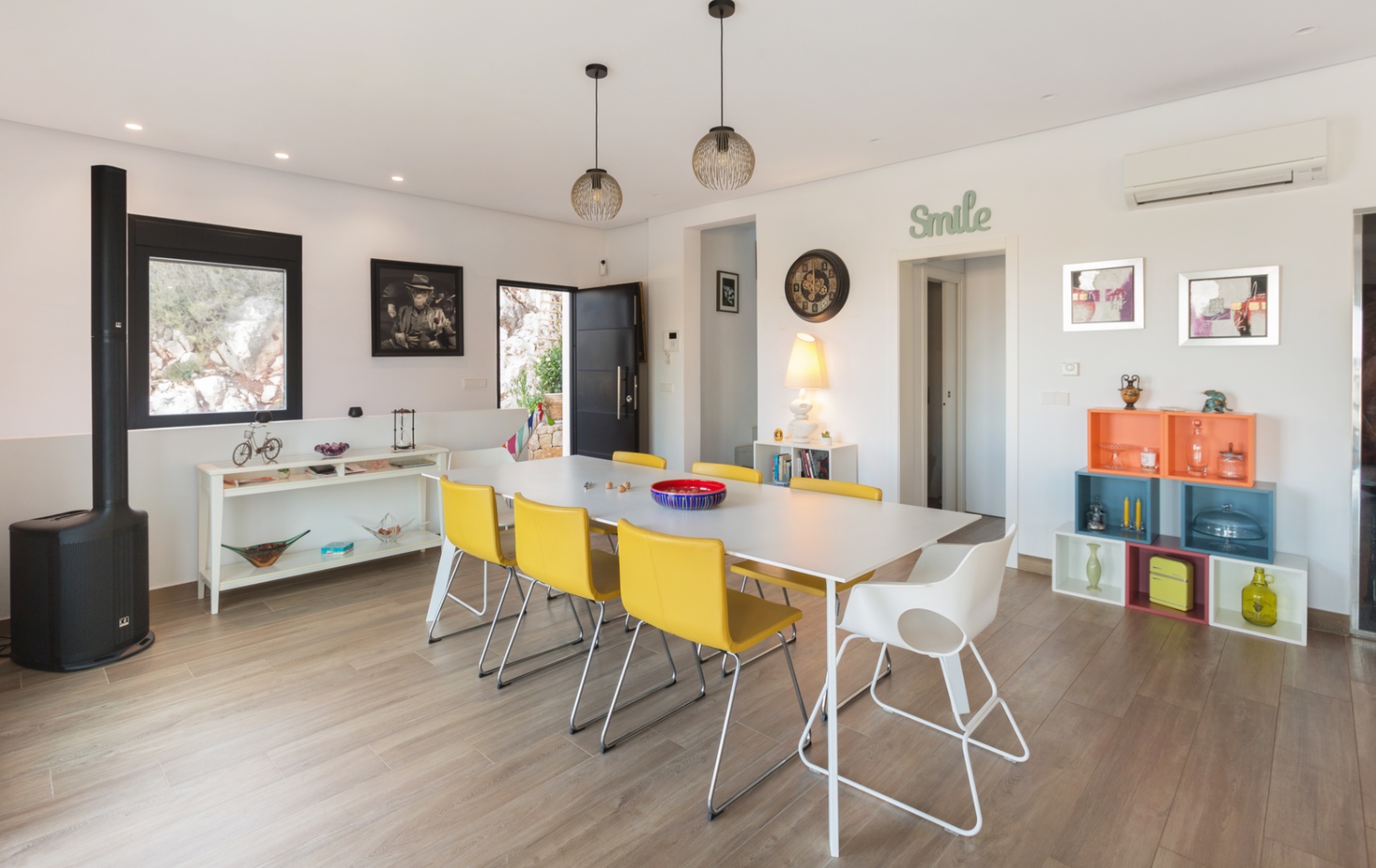 Luminoso comedor con mesa blanca, seis sillas amarillas y una blanca, bajo lámparas colgantes modernas. Destaca el suelo de imitación madera y decoración ecléctica. Luminoso comedor con mesa blanca, seis sillas amarillas y una blanca, bajo lámparas colgantes modernas. Destaca el suelo de imitación madera y decoración ecléctica.