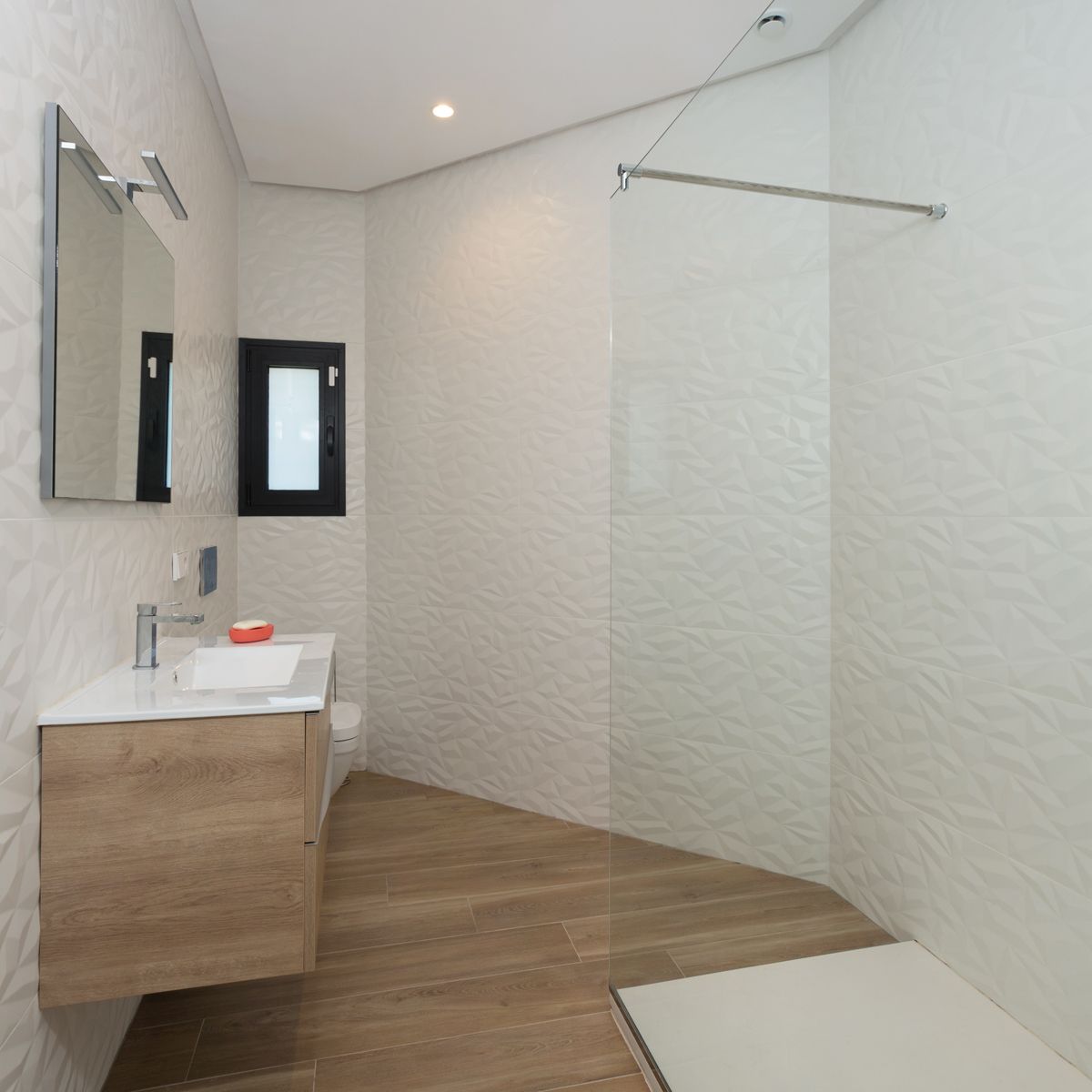 Moderno baño con mueble de madera suspendido, lavabo blanco, espejo iluminado y ducha de obra con mampara de cristal. Destacan los azulejos blancos con textura geométrica y el suelo de imitación madera. Moderno baño con mueble de madera suspendido, lavabo blanco, espejo iluminado y ducha de obra con mampara de cristal. Destacan los azulejos blancos con textura geométrica y el suelo de imitación madera.