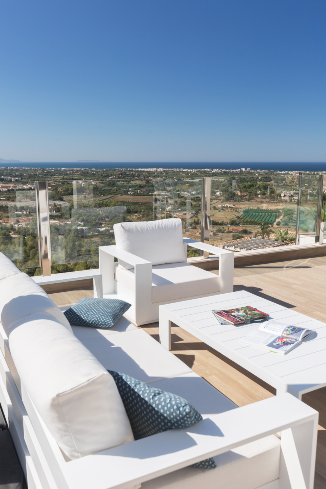 Zona de estar exterior con mobiliario blanco moderno sobre tarima de madera, barandilla de cristal y vistas panorámicas a la costa y al mar Mediterráneo bajo un cielo despejado. Zona de estar exterior con mobiliario blanco moderno sobre tarima de madera, barandilla de cristal y vistas panorámicas a la costa y al mar Mediterráneo bajo un cielo despejado.