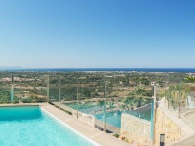 Amplia terraza exterior con piscina rectangular, mobiliario moderno blanco para comedor y salón, y tumbonas. Vistas panorámicas a la costa, el mar y las colinas verdes bajo un cielo despejado.