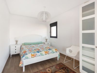 Dormitorio con cabecero de lamas blanco, edredón colorido y suelo de baldosas imitación madera. Incluye mesitas de noche blancas con lámparas decorativas, ventana de marco oscuro y armario con paneles de cristal esmerilado.