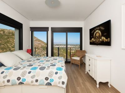 Dormitorio luminoso con cama de diseño, suelo de madera y amplios ventanales negros que enmarcan vistas panorámicas a la montaña y la costa. Incluye cómoda blanca y TV.