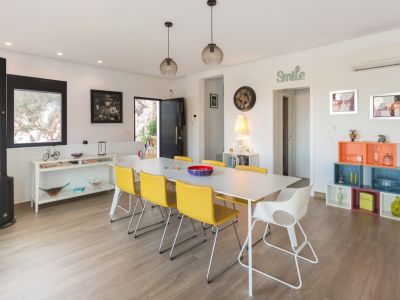 Luminoso comedor con mesa blanca, seis sillas amarillas y una blanca, bajo lámparas colgantes modernas. Destaca el suelo de imitación madera y decoración ecléctica.