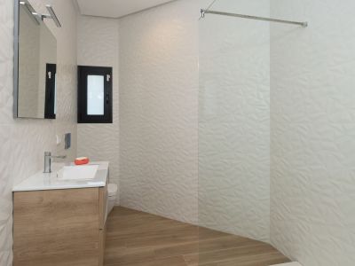 Moderno baño con mueble de madera suspendido, lavabo blanco, espejo iluminado y ducha de obra con mampara de cristal. Destacan los azulejos blancos con textura geométrica y el suelo de imitación madera.