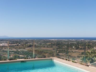 Piscina de borde infinito con agua azul cristalina y barandilla de cristal, ofreciendo vistas panorámicas al paisaje costero y al mar Mediterráneo bajo un cielo despejado.