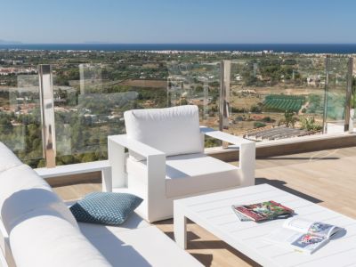 Zona de estar exterior con mobiliario blanco moderno sobre tarima de madera, barandilla de cristal y vistas panorámicas a la costa y al mar Mediterráneo bajo un cielo despejado.
