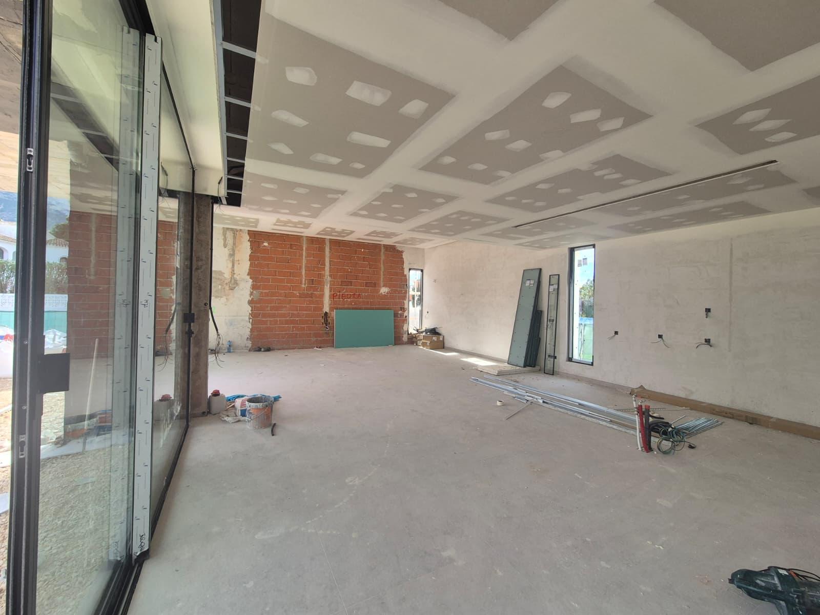 Amplio salón en construcción con techo de pladur preparado para iluminación, pared de ladrillo visto y grandes ventanales correderos de suelo a techo.