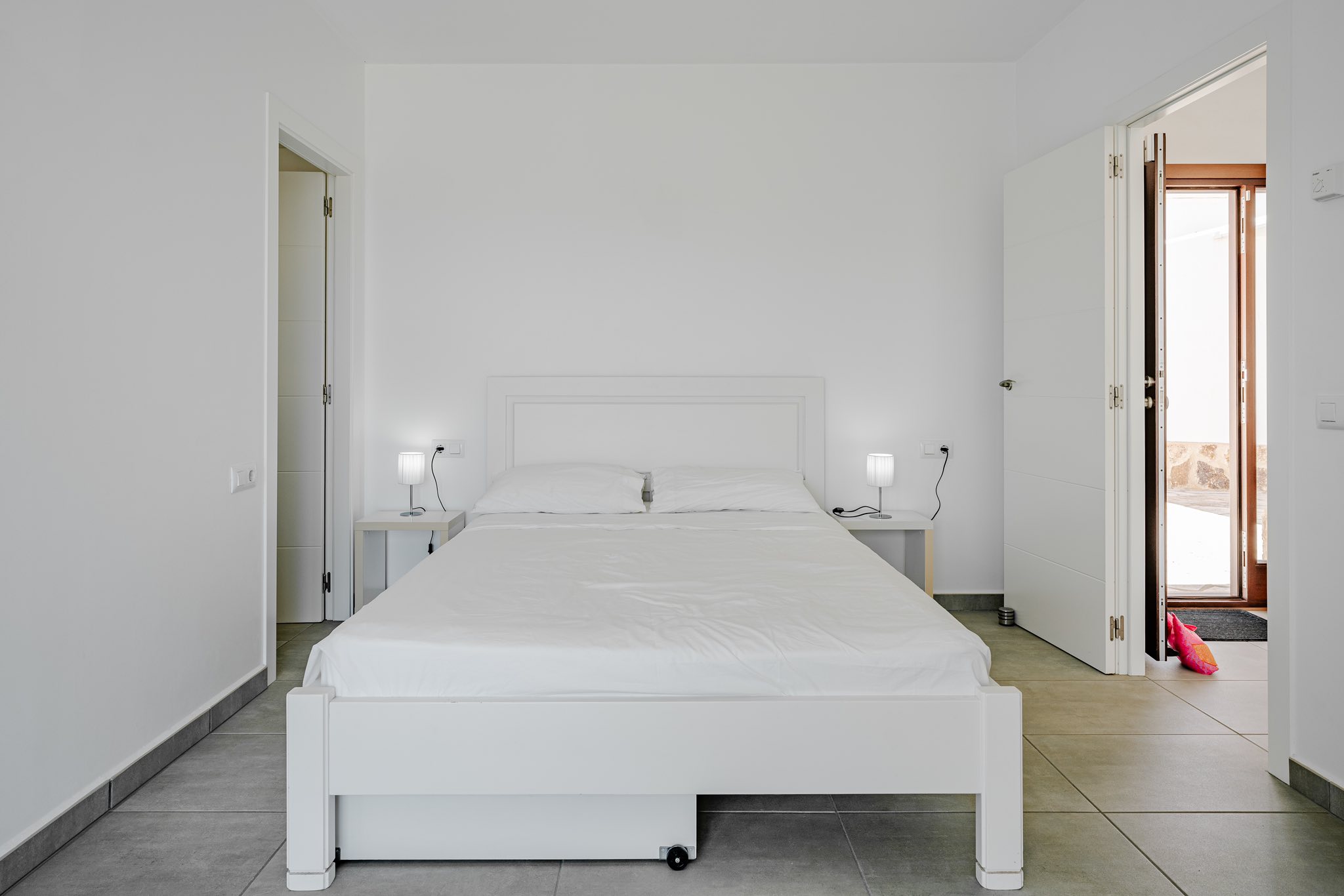 Dormitorio minimalista con cama de madera blanca, mesitas de noche a juego y suelo de baldosas grises. Las puertas abiertas conectan con el exterior y otras estancias.