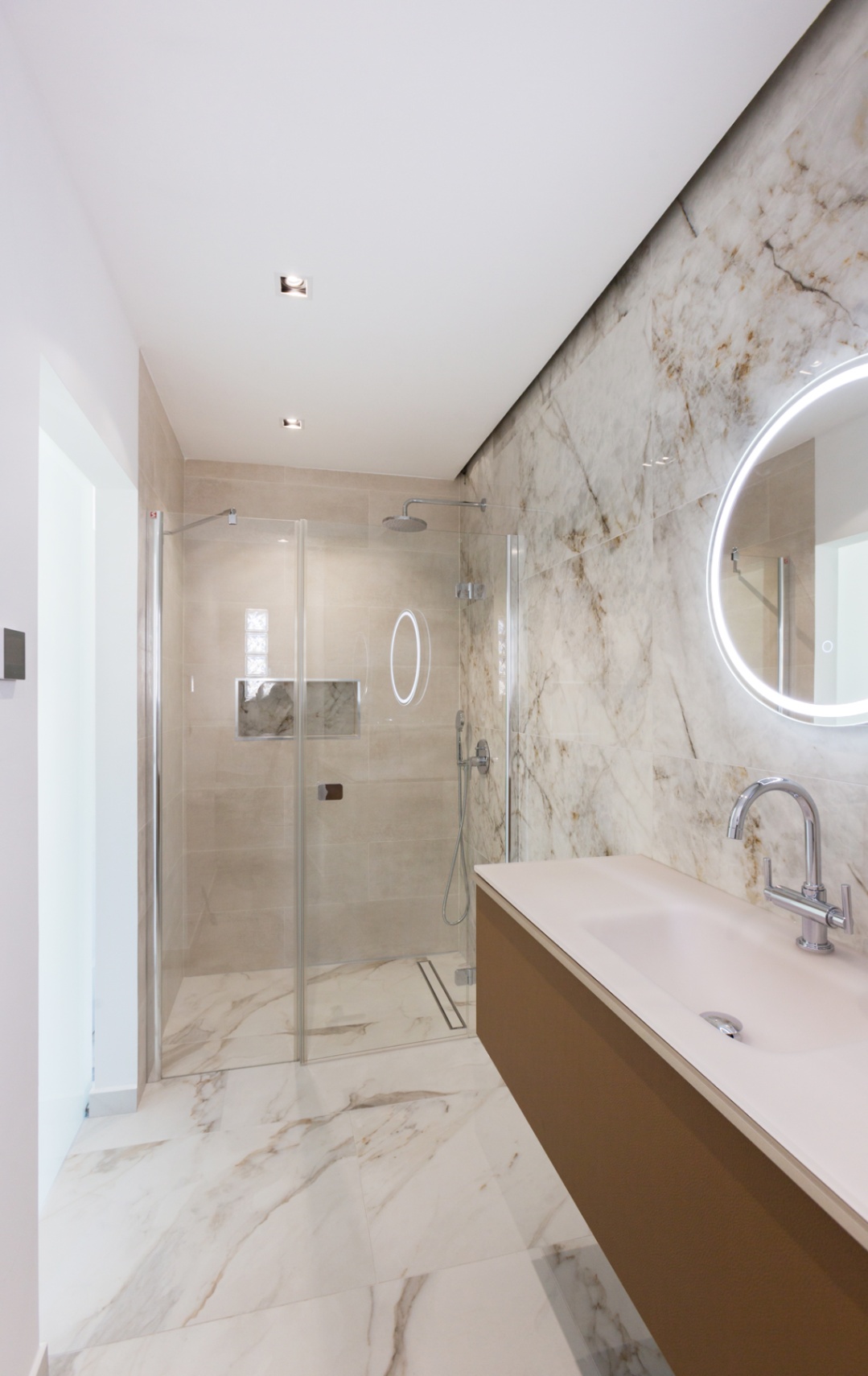Baño moderno con ducha de obra acristalada, revestimiento de mármol veteado, mueble suspendido con lavabo integrado y espejo circular con retroiluminación LED.