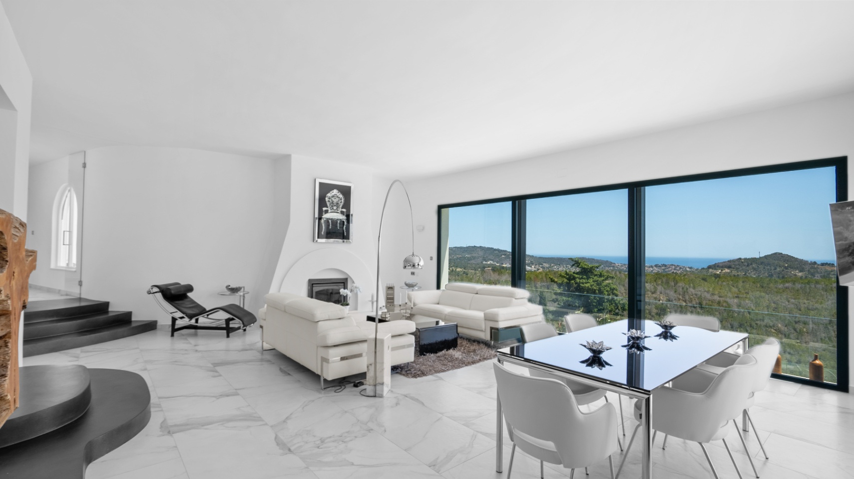 Salón comedor de diseño abierto con suelos de mármol blanco, chimenea integrada y amplios ventanales con vistas panorámicas al valle y al mar Mediterráneo.
