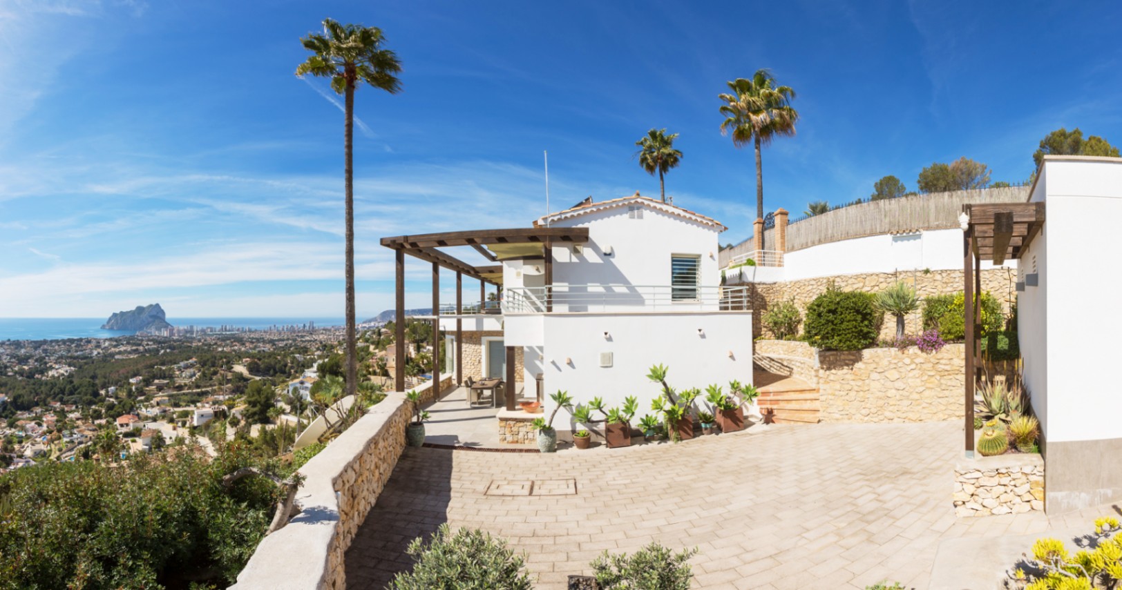Exterior de una villa mediterránea con fachadas blancas, pérgolas de madera y muros de piedra, con vistas panorámicas a la costa y al Peñón de Ifach.