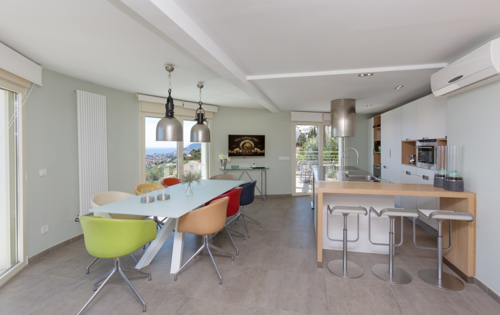 Interior diáfano con cocina moderna, barra de desayuno de madera y comedor con sillas coloridas y lámparas industriales, con amplios ventanales hacia la costa.