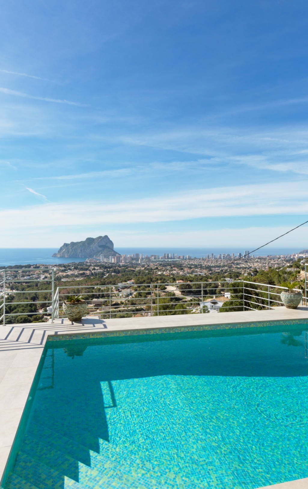 Terraza elevada con piscina de mosaico turquesa y borde de piedra blanca, con vistas panorámicas al Peñón de Ifach y la costa mediterránea bajo un cielo despejado.