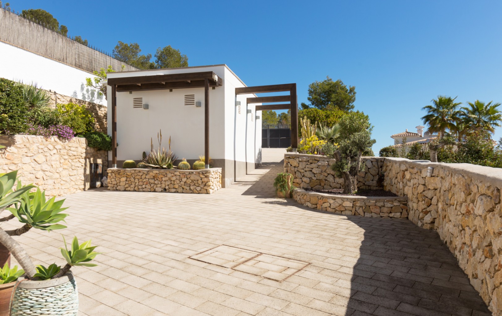 Zona exterior pavimentada con muros de piedra seca, jardineras con cactus y una estructura blanca con vigas de madera oscura bajo un cielo despejado.
