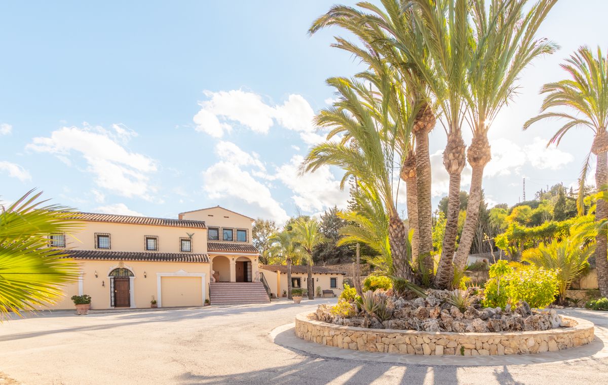 Amplio camino de entrada pavimentado con una jardinera circular de piedra y palmeras. La villa presenta fachadas en tonos crema y cubiertas de teja árabe.