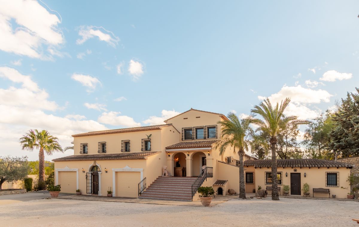Fachada exterior de una villa mediterránea con tejado de terracota, amplia escalinata hacia el porche arqueado, garajes integrados y palmeras en el acceso.